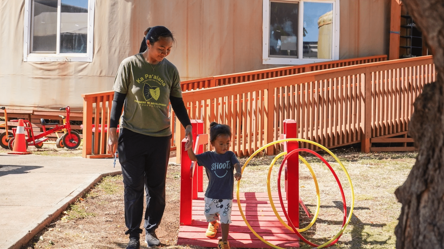 Caregiver guides a young child through a small outdoor obstacle course with red rails and yellow hoops outside a building.