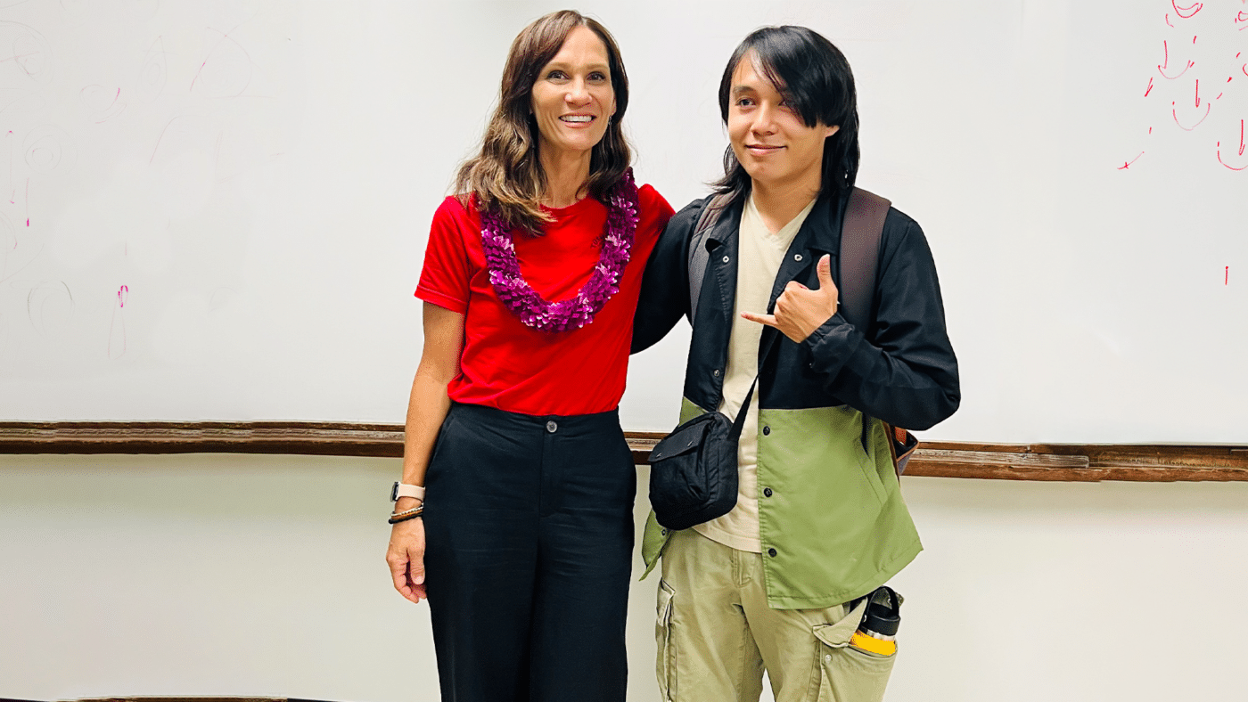 Two people posing in a classroom: a woman in a red shirt with a purple lei and a man in a beige/green outdoor jacket smiling.